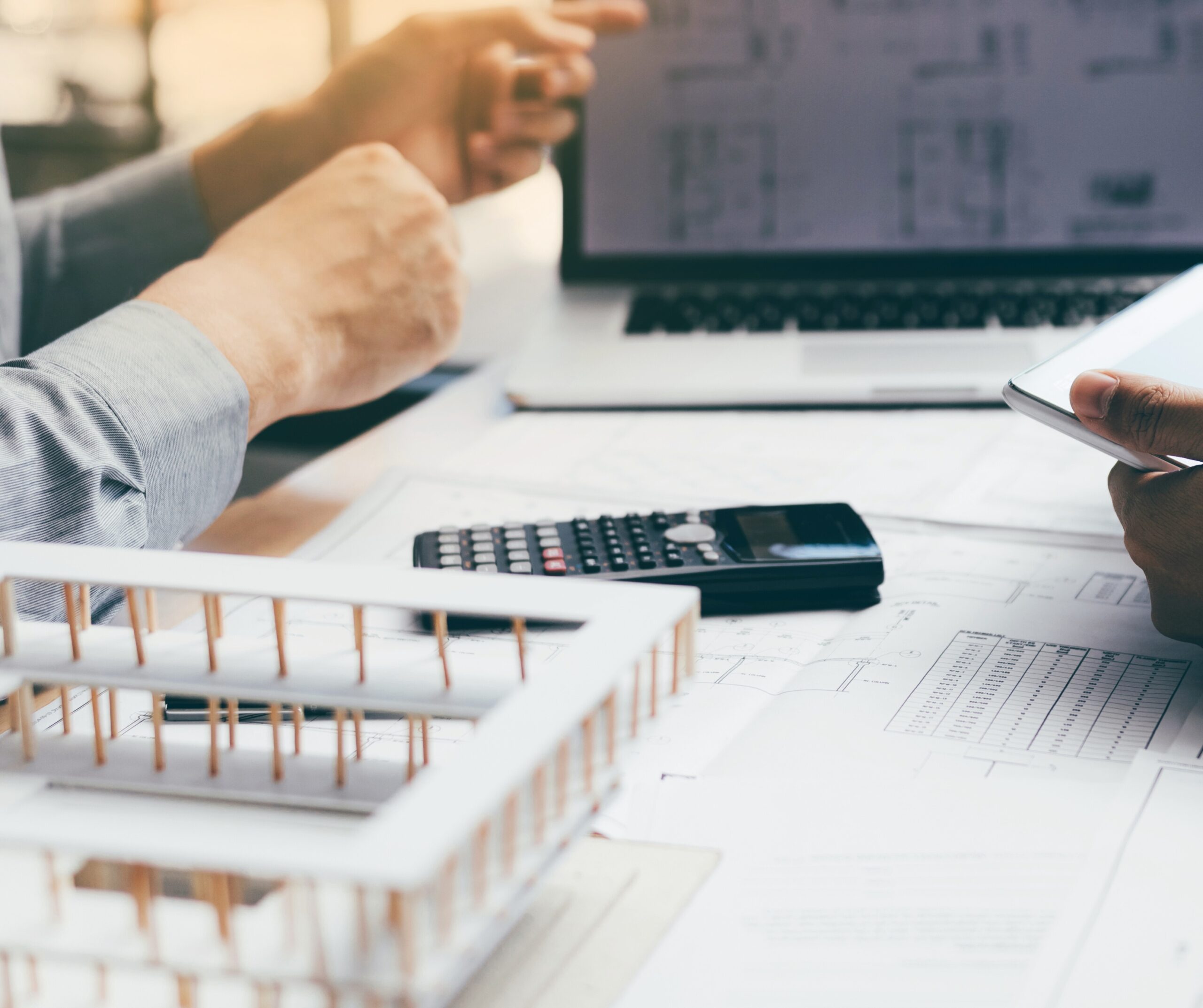 Two professionals reviewing architectural plans and project financials on a tablet and laptop, with a calculator and a building model in view, illustrating WIP accounting and financial management in construction.