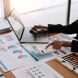 Business professional reviewing financial reports with a laptop and calculator, surrounded by charts and graphs showing business performance metrics.