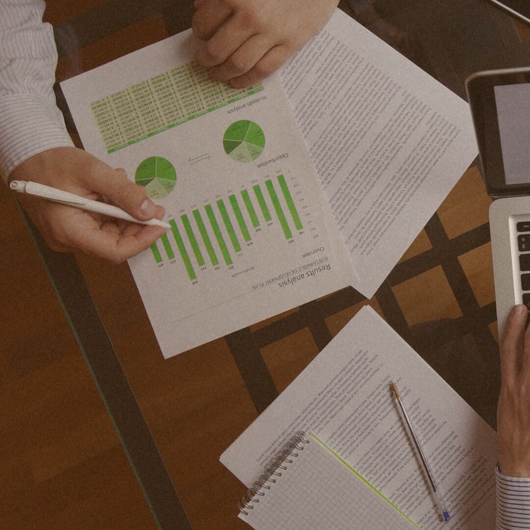 Top-down view of hands analyzing business reports with green charts and a laptop on a glass desk.