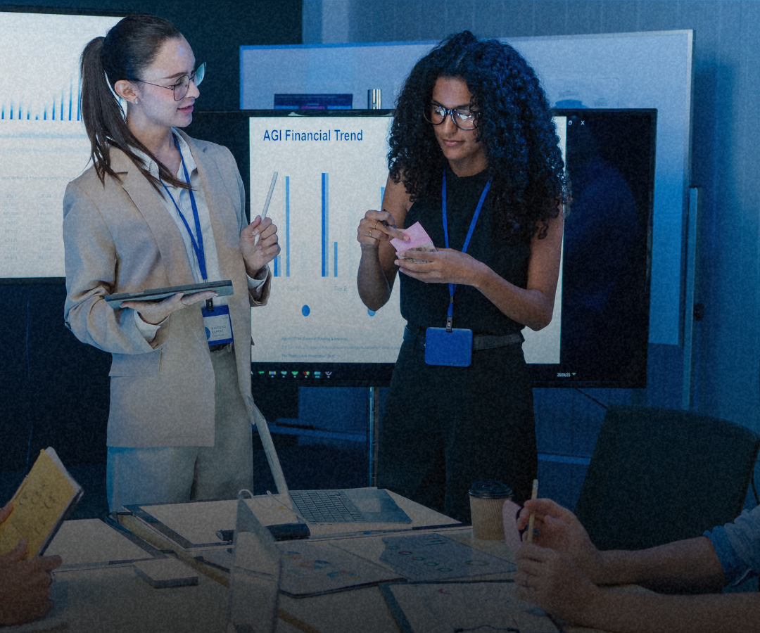 How Often Should You Analyze Where Your Business Stands? Two colleagues review financial data during a meeting, with a bar chart showing financial trends displayed on a monitor behind them, while others take notes at the table.